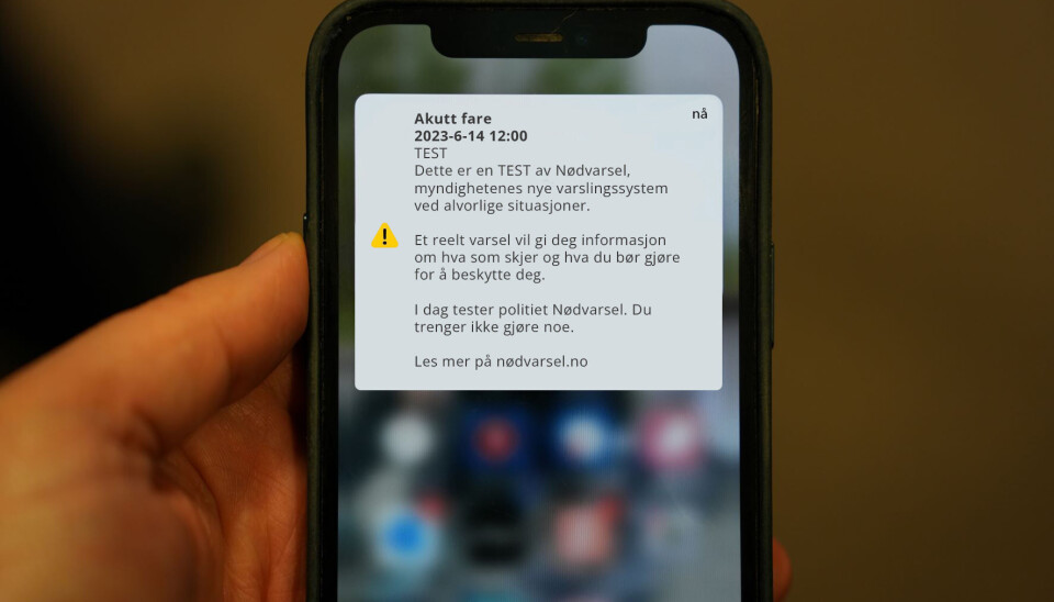 Bildet er av en hånd som holder en mobiltelefon. En firkant med informasjon om 'akutt fare' lyser opp foran alt annet på skjermen. Det er et eksempel på hvordan det nasjonale nødvarslet skal se ut. Meldingen blir også lest opp for folk som har 'tekst-til-tale' aktivert på sin mobil.