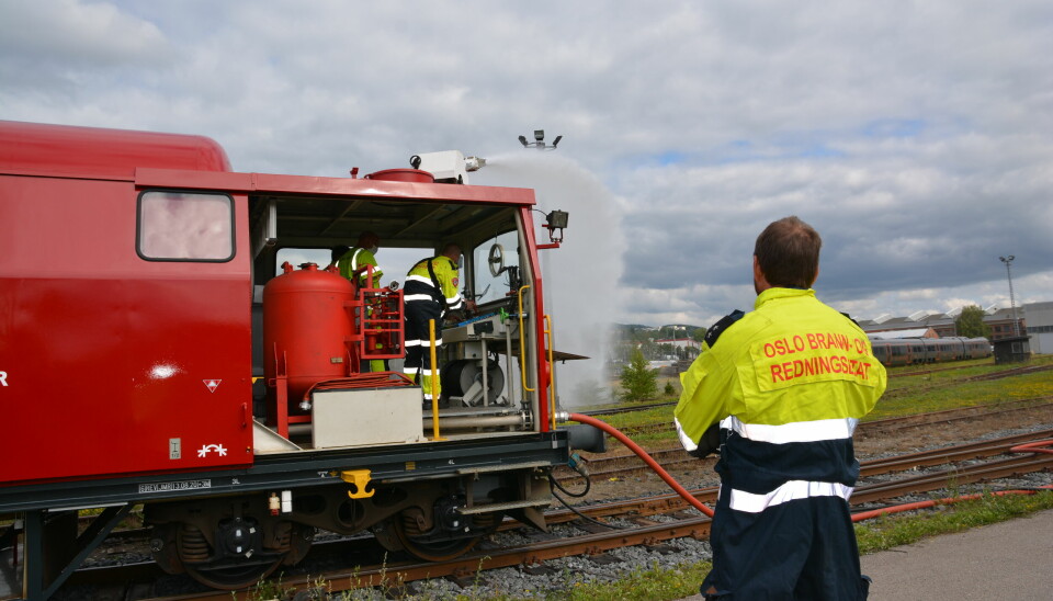 Bildet er av lokomotivet på brann- og redningstoget. En kraftig vannstråle kommer ut av vannkanonen på taket av lokomotivet. En mann i arbeidsuniform fra Oslo brann- og redningsetat ser på. Det er også slangeutlegg koblet på fronten av toget.