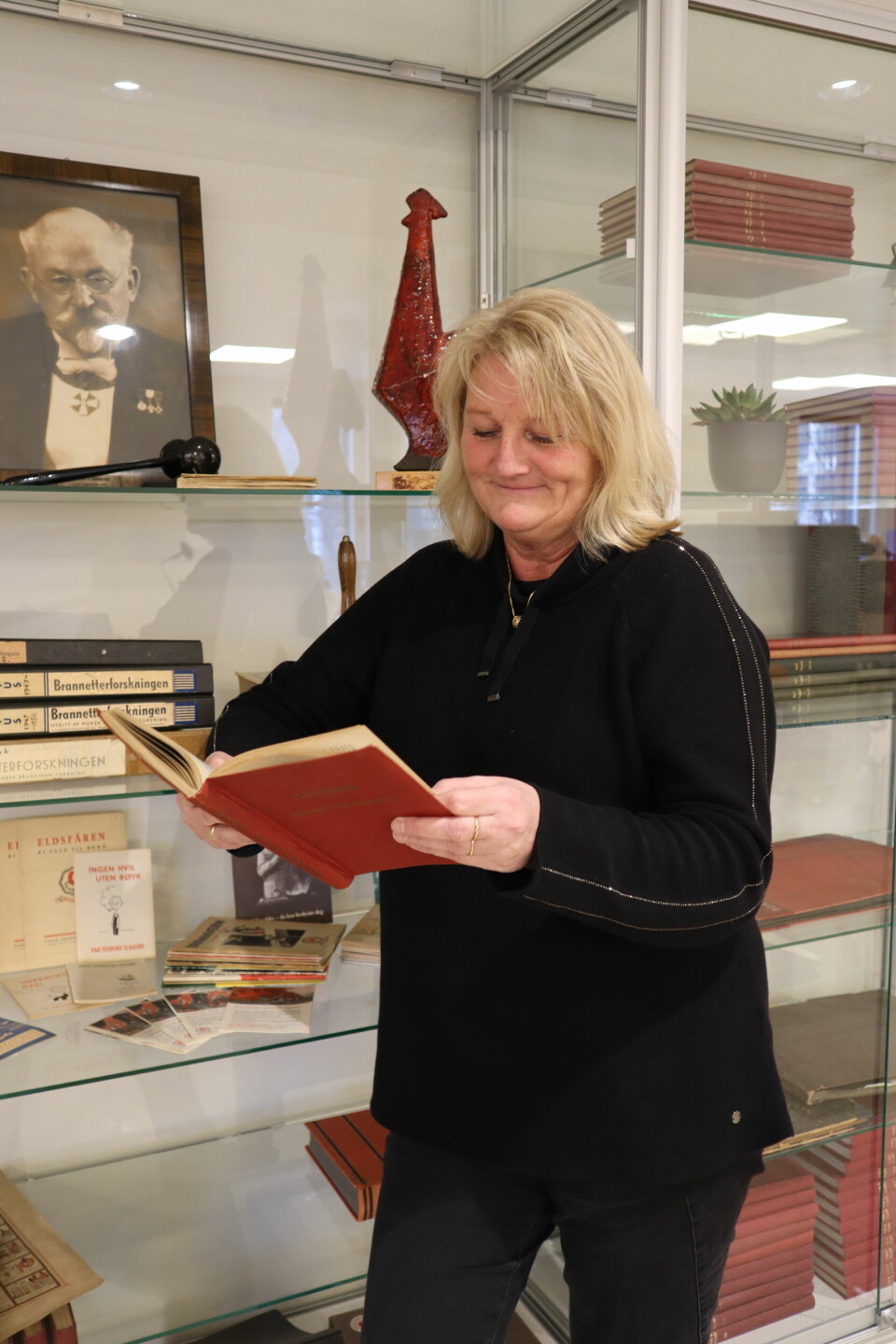 Bildet er av Sølvi M. Harjo som blar i en bok. Hun står foran en glassmonter med historiske bøker, bilder og gjenstander fra Brannvernforeningen.