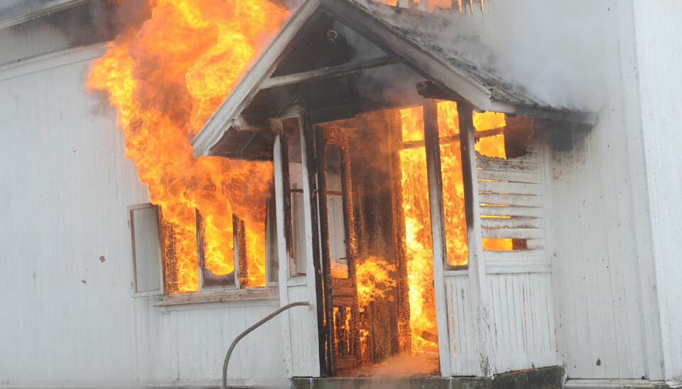 Bildet er et illustrasjonsfoto av et hvitt hus som brenner. Flammene står ut av både inngangsdøra, et vindu og fra veggen i andreetasje.