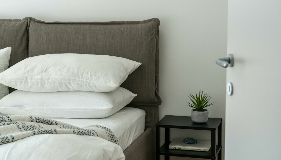 Bildet er et utsnitt fra et soverom. Bildet er tatt gjennom en dør, og viser en delvis oppredd seng og et lite nattbord med en bok og plante.